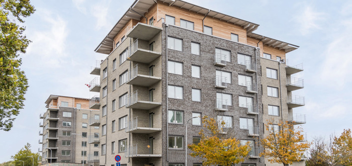 Flerbostadshus i grått tegel med balkonger och träfasad högst upp, sett från gatan med höstfärgade träd framför.